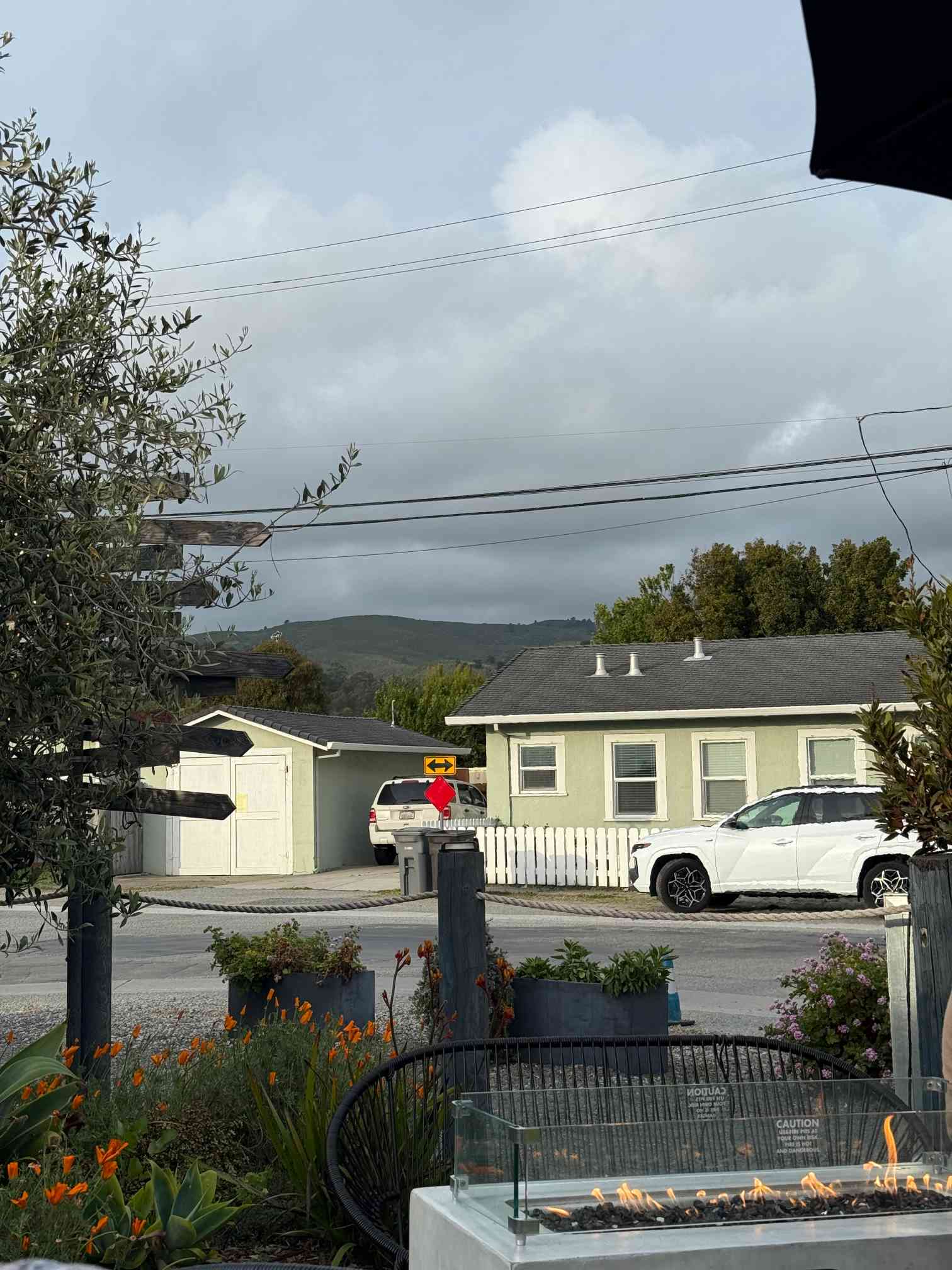 Residential scene with a cream-colored house with gray roof, white picket fence, and two white vehicles parked nearby. In the foreground is a glass-enclosed fire table and orange flowers. Background shows hills under an overcast sky with power lines crossing above. A person in red stands between vehicles near a yellow directional sign.
